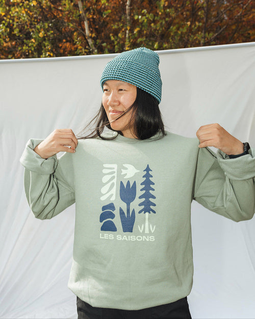 person wears a sage hooded crewneck with front graphic from the brand les Saisons