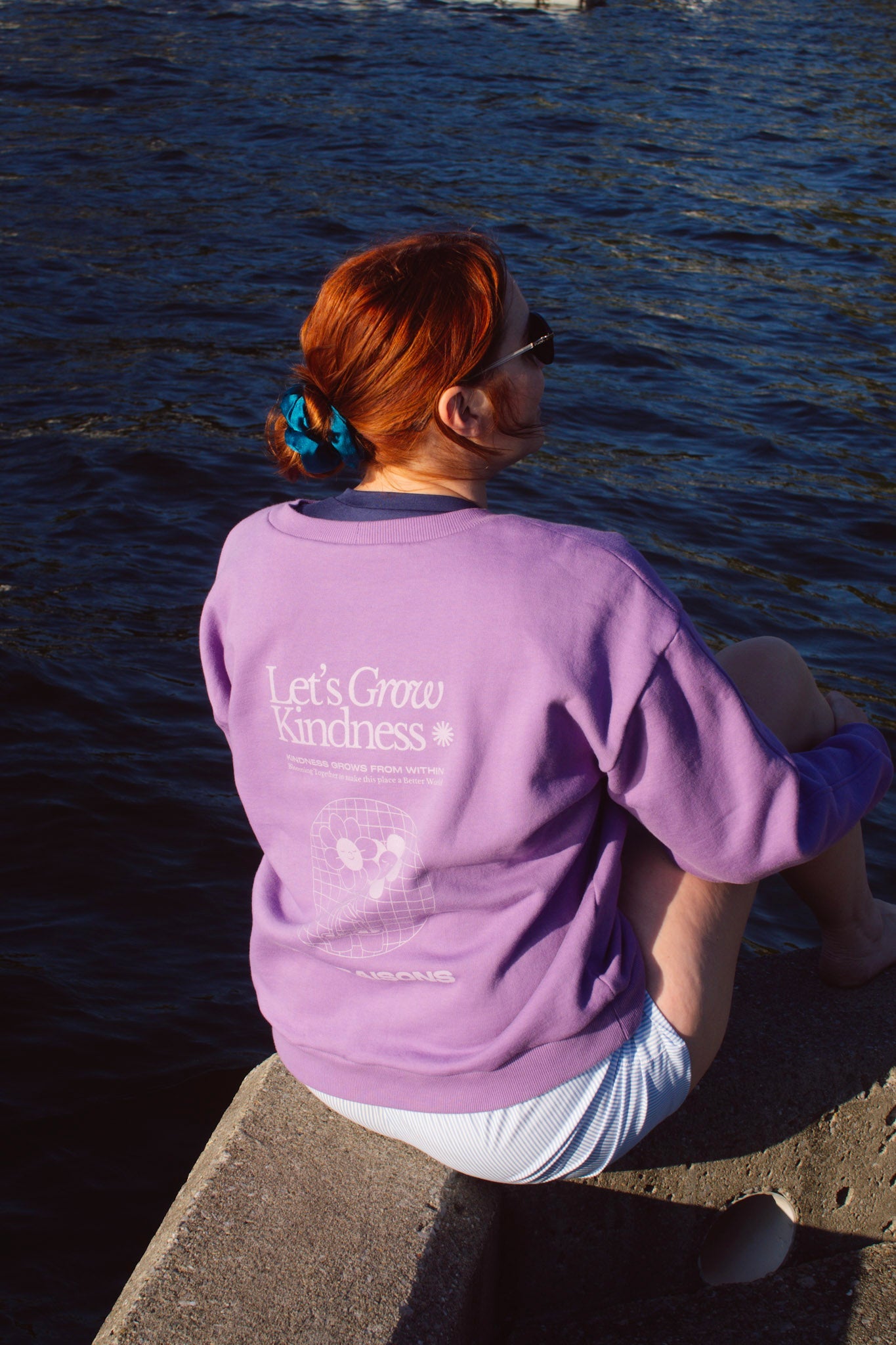 Person wearing a purple sweatshirt graphic with growing kindness by a body of water, crewneck made in canada
