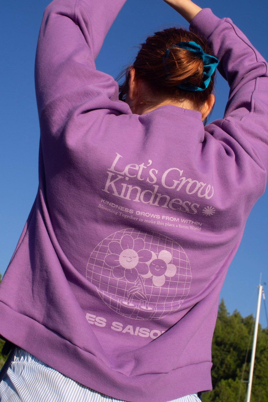 Person wearing a purple hoodie with 'Let's Grow Kindness' text and design, standing outdoors against a clear blue sky.