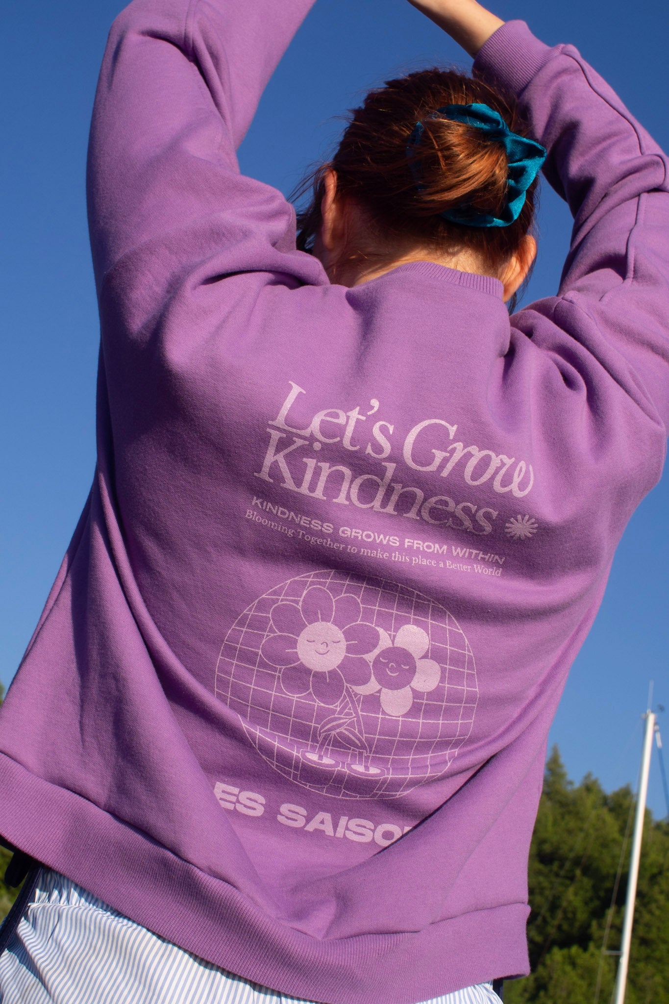Person wearing a purple hoodie with 'Let's Grow Kindness' text and design, standing outdoors against a clear blue sky.