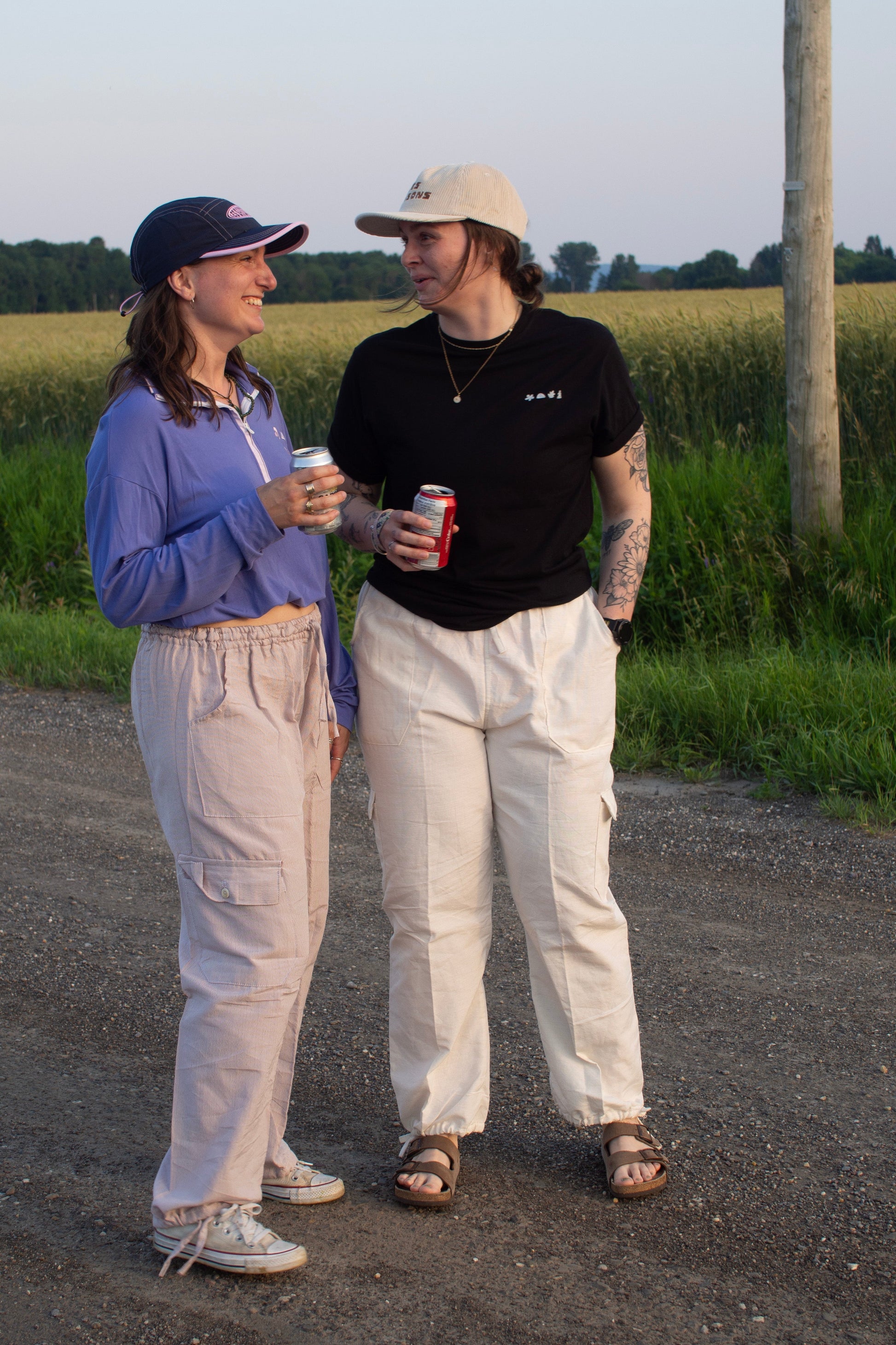 Two people standing on a road near a field, holding drinks and smiling.