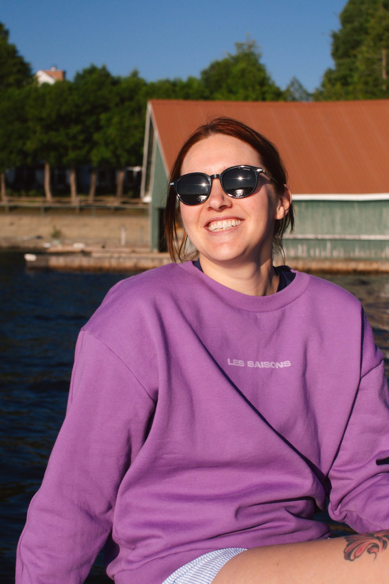 Person wearing a purple sweatshirt with sunglasses, sitting by a body of water with a cabin in the background.